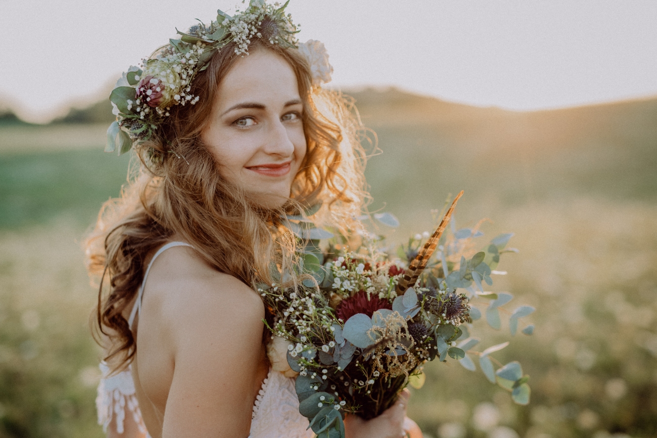 Beautiful young bride with flower wreath outside in green nature at sunset.